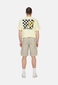 Camiseta de algodón amarillo claro con un logo de tablero de ajedrez en blanco y negro y texto amarillo que dice "MTV MUSIC TELEVISION", combinada con pantalones cortos cargo grises.