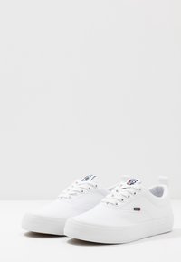 Baskets en toile blanche avec bout rond, surface texturée, semelle en caoutchouc plate et languette marquée featuring des accents rouges et bleus.