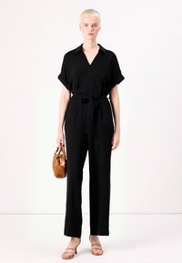 Svart jumpsuit med kort ermer, omslagstype foran, belte i midjen, vide ben og glatt tekstur; sammen med en brun håndveske.