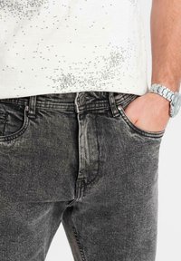 Jeans de mezclilla negros con una textura deslavada, diseño de cinco bolsillos y herrajes metálicos. Combinados con una camisa blanca de patrón gráfico.