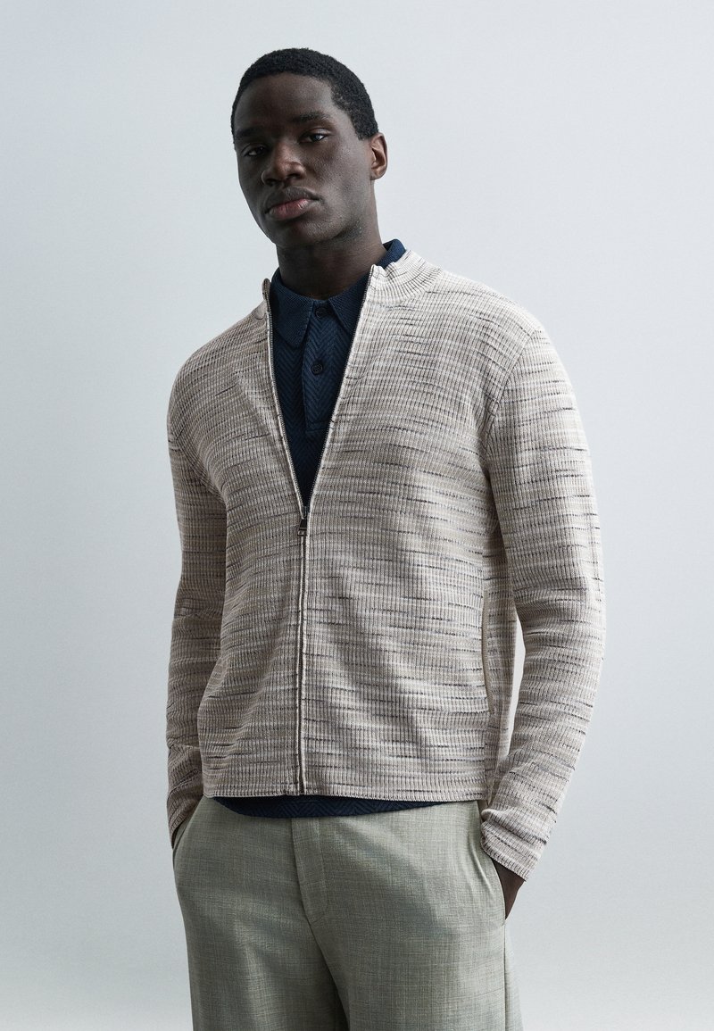 Homme portant une veste zippée texturée beige clair sur une chemise bleu marine et un pantalon gris clair, debout les mains dans les poches devant un fond uni.