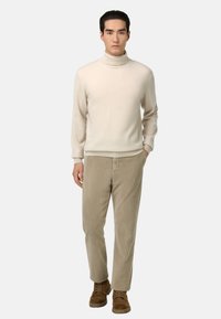Un pull à col roulé crème avec des poignets côtelés, associé à un pantalon en velours côtelé beige et des chaussures en daim marron, se tenant contre un fond gris.