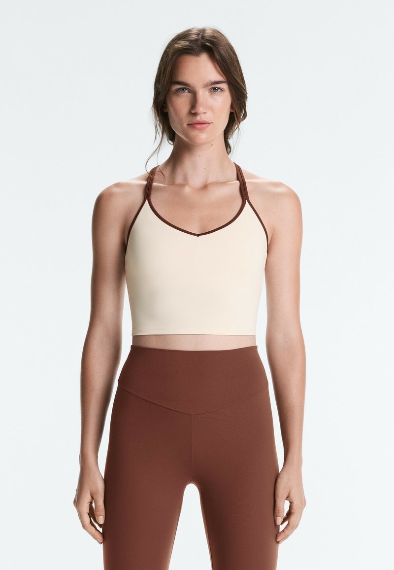 Femme portant un débardeur court beige avec un liseré marron et un legging marron taille haute, debout devant un fond blanc uni.