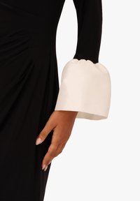 Czarna odzież o dopasowanej sylwetce, z białym satynowym mankietem w kształcie dzwonu z marszczeniem. Gładka tekstura, elegancki kontrast kolorystyczny.