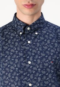 Chemise à manches longues bleu marine avec un motif cachemire blanc, col boutonné et boutons blancs ; elle présente un petit logo rouge et blanc sur la poitrine.