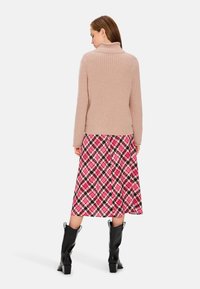 Beige gestrickter Rollkragenpullover mit gerippter Textur kombiniert mit einem rosa und schwarzen Rock im Rautenmuster, ergänzt durch schwarze Stiefeletten.