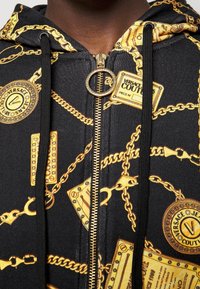 Hoodie zippé noir avec un motif de chaînes et de logos dorés, doté d'une fermeture éclair métallique et d'un tirette en anneau sur le devant.