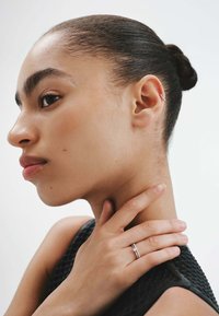 Anillo de plata que se lleva en el dedo índice, con un diseño simple. El tono de piel de la persona es natural y lleva una prenda negra texturizada.