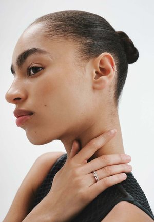 Anillo de plata que se lleva en el dedo índice, con un diseño simple. El tono de piel de la persona es natural y lleva una prenda negra texturizada.