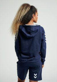 Marinblå huvtröja med grå insida, med vita chevron detaljer på ärmarna och en avslappnad passform, tillsammans med marinblå shorts.