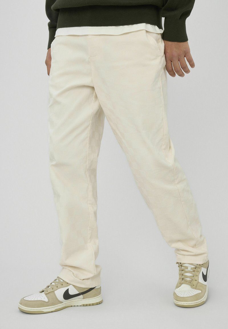 Pantalon en velours côtelé beige clair avec une texture côtelée verticale, associé à des baskets Nike beige et blanches avec des accents noirs.