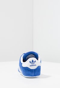 Modrá semišová teniska s bílými detaily, kulatým podpatkem, polstrovaným lemem a logem Adidas na zadní straně. Hladká podrážka pro lepší trakci.