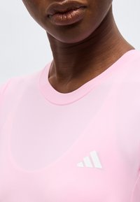 Κοντινό πλάνο του κάτω μέρους του προσώπου και του λαιμού ενός ατόμου που φορά ένα ανοιχτό ροζ πουκάμισο με μικρό λευκό λογότυπο Adidas στο στήθος.