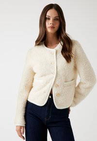 Giovane donna che indossa una giacca a bottoni di un colore crema con textura e tasche frontali, abbinata a jeans a vita alta blu scuro, su sfondo bianco.