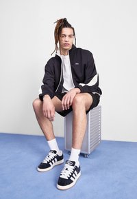 Svart zip-up jacka med krämfärgade detaljer, grå t-shirt, svarta shorts och svarta mockasneakers med vita ränder. Sitter på en metallpall.