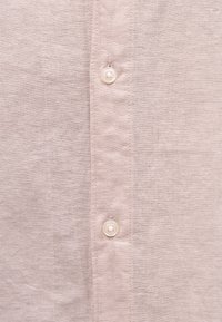 Ελαφρύ μπεζ ύφασμα με δύο μικρά λευκά κουμπιά ραμμένα στο κέντρο, που δείχνουν λεπτομερή υφή και ραφές του μπροστινού πάνελ ενός πουκαμίσου.