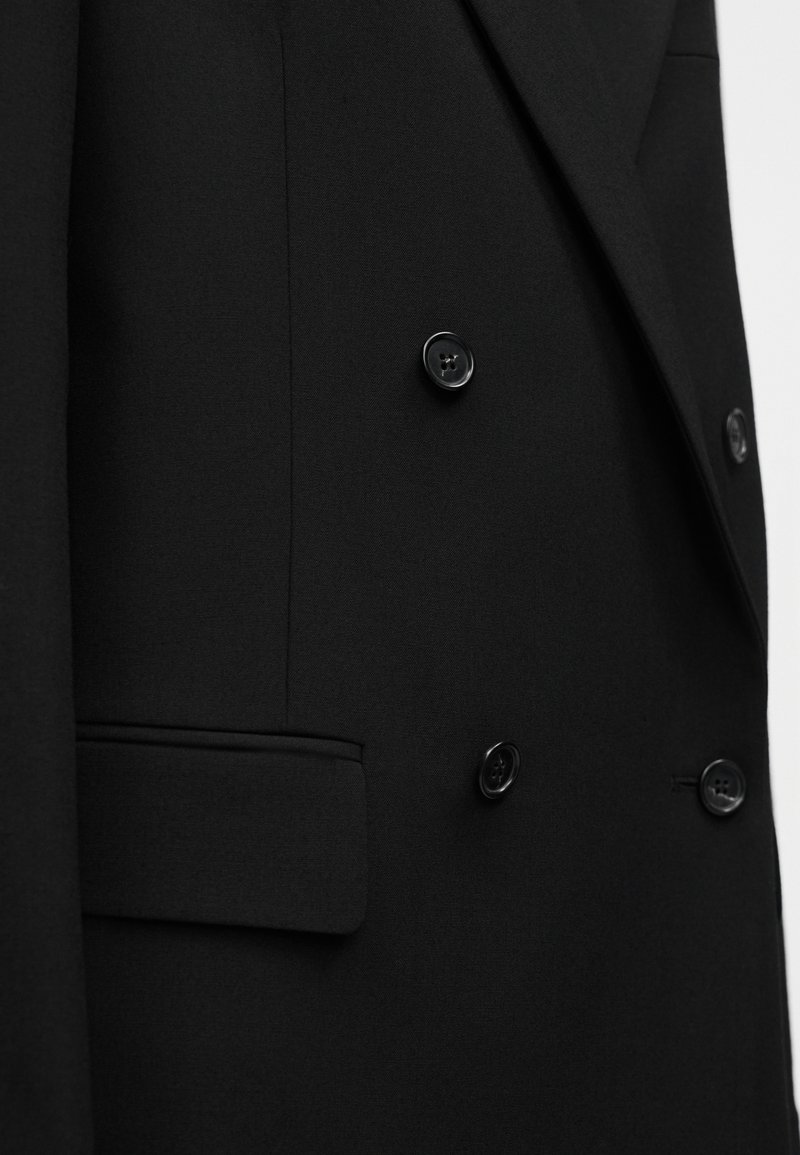 Blazer noir croisé à double boutonnage, en tissu lisse, coupe slim, deux boutons, et poche latérale avec une texture subtile.