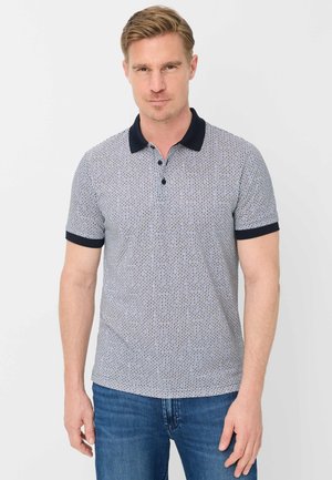 Uomo biondo che indossa una polo a maniche corte blu navy e bianca con motivi, e jeans blu, in piedi davanti a uno sfondo chiaro e uniforme.