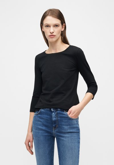Femme aux longs cheveux bruns portant un t-shirt noir à manches longues et un jean bleu, debout avec une main dans la poche, sur un fond uni.