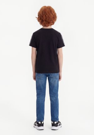 Bambino con capelli rossi ricci che indossa una t-shirt nera, jeans blu e sneakers nere e bianche, in piedi di spalle su sfondo bianco.