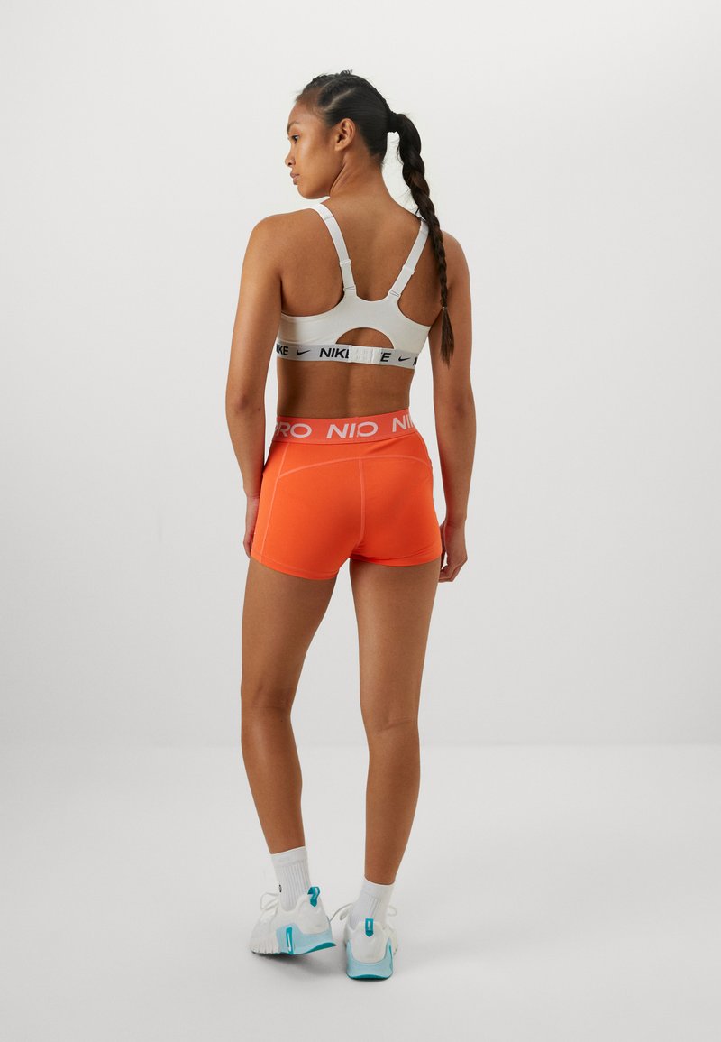 Sujeción deportiva en gris claro con tirantes ajustables y detalle cortado. Pantalones cortos naranjas con cinturilla elástica blanca. Zapatillas deportivas blancas con acentos azules.