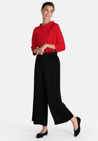 Maglia rossa a maniche lunghe con scollo a cowl, abbinata a pantaloni larghi neri e scarpe basse nere. Accessori in argento semplici inclusi.