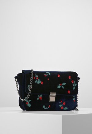 Kleine donkerblauwe fluwelen schoudertas met rood en groen bloemrijk borduurwerk, een zilveren kettingriem en sluiting, tentoongesteld op een witte ondergrond.