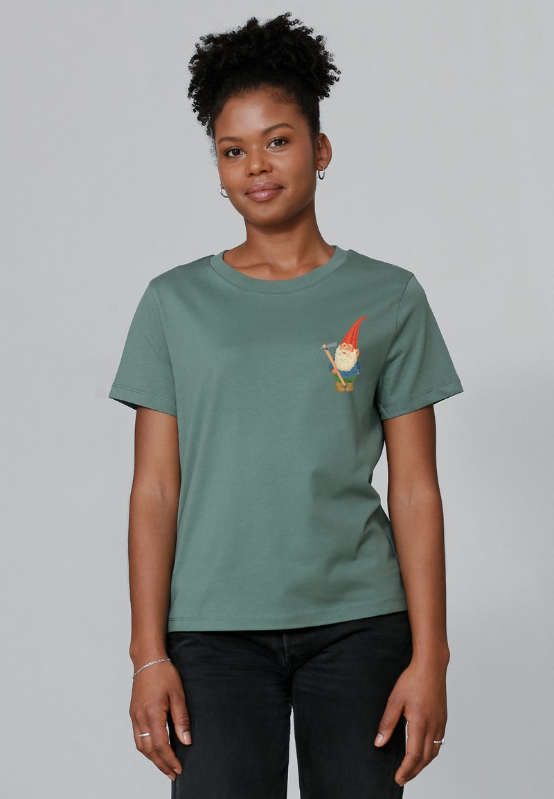 T-shirt en coton vert avec un graphique d'un gnome portant un chapeau rouge sur la poitrine gauche. Manches courtes et encolure ras du cou, porté avec un pantalon noir.