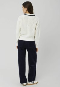 Pull en laine blanc avec un motif texturé et un col marine, associé à un pantalon en velours côtelé bleu foncé et des chaussures blanches avec des accents noirs.