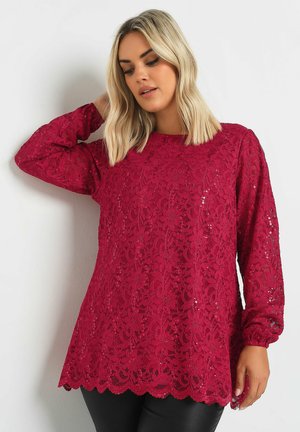 Top de encaje rojo con mangas largas, dobladillo con ondas y lentejuelas. Presenta un escote redondo y patrones florales intrincados en una tela texturizada.