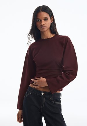 Junge Frau mit langen dunklen Haaren, die ein bordeauxrotes Langarmshirt und dunkelblaue Jeans vor einem schlichten weißen Hintergrund trägt.