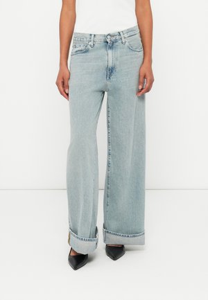 Széles szárú világoskék farmernadrág, magas derékkal, felgöngyölt szegéllyel és elülső zsebekkel, fekete hegyes orrú cipővel viselve.