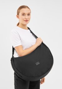 Borsa a spalla in tessuto nero con forma arrotondata, tracolla regolabile e una evidente patch con logo. Texture liscia, design minimalista.
