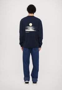 Námořnická modrá mikina s kulatým výstřihem, na zadní straně s grafikou slunce a vlny. Pod designem je vytištěn text. Nosená s modrými džíny.