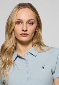 Ljusblå polo skjorta i texturerad tyg, med krage, knappstängning och en liten broderad logotyp på bröstet.