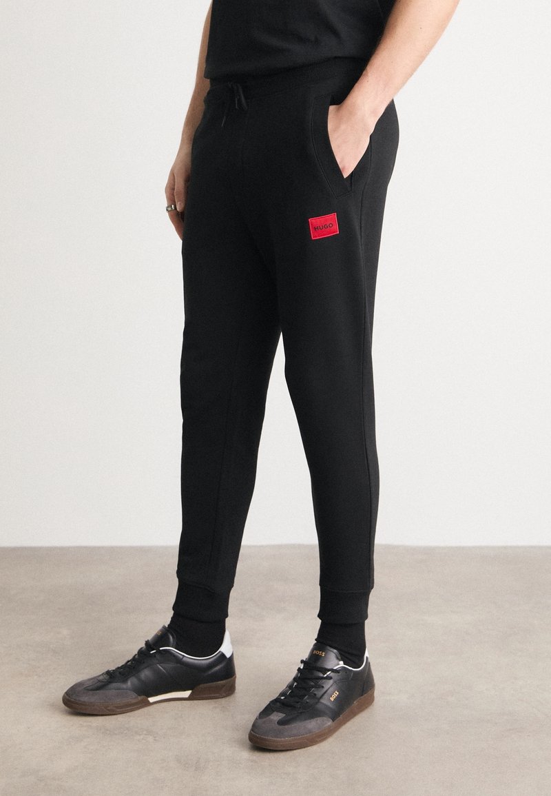 Persona vestida con pantalones de chándal negros con parche rojo del "Hugo", zapatillas negras con suelas marrones, de pie sobre un suelo gris frente a una pared blanca.