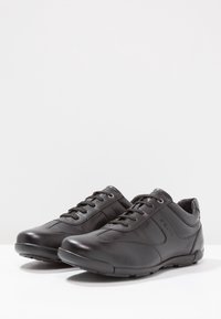 Chaussures de sport en cuir noir avec bout arrondi, lacets à l'avant, texture lisse et marquage discret sur le côté. Semelles en caoutchouc pour une bonne adhérence.