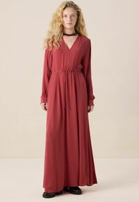 Femme avec de longs cheveux blonds ondulés portant une robe rouille longue avec une ceinture à la taille et de grosses chaussures noires, sur un fond uni.