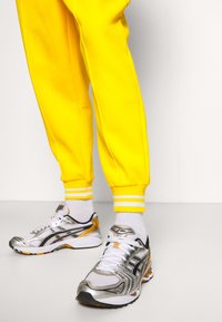 Silberne und schwarze Sportschuhe mit weißem Mesh, einer gepolsterten Sohle und einem orangefarbenen Akzent. Kombiniert mit gelben Jogginghosen, die weiße Streifen haben.