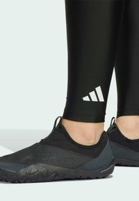 Schwarzer Sportschuh mit einem strukturierten Mesh-Obermaterial, das ein graues Akzentdetail und ein auffälliges Logo aufweist. Getragen mit schwarzen, enganliegenden Leggings, die ein weißes Logo zeigen.