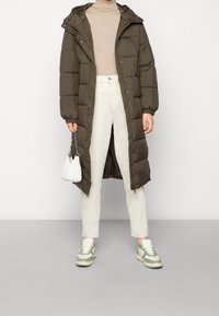 Pehmustettu oliivinvihreä pitkä takki hupulla, sivutaskuilla ja napeilla, puettuna beigenvärisen ribbineuleen ja vaaleiden housujen päälle. Valkoinen käsilaukku ja lenkkarit.