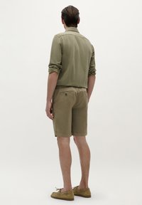 Căma�șă verde măsliniu cu mâneci suflate, combinată cu pantaloni scurți bej cu buzunare la spate și pantofi casual maro deschis.