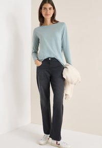 Maglietta a maniche lunghe azzurro chiaro abbinata a pantaloni di jeans scuri e sneakers bianche. Un maglione crema è tenuto nella mano sinistra.