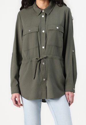 Femme portant une chemise boutonnée à manches longues vert olive avec poches poitrine et taille coulissante, associée à un jean bleu clair.