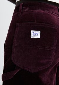 Gros plan sur la poche arrière d'un pantalon en velours côtelé violet foncé avec une étiquette blanche Lee Workwear et des passants de ceinture visibles.