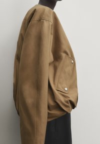 Veste en coton marron oversize avec un col rond et une poche plaquée. Présente des accessoires en métal argenté et des détails côtelés sur les manches.