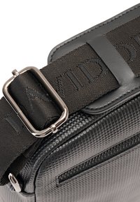 Zwarte textuur tas met een geometrisch patroon, voorzien van een zilveren metalen gesp en een dikke zwarte strap met reliëfletters.