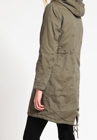 Olivgrön parkasjacka med huva, med dragsko i midjan, framfickor och ett lätt strukturerat tyg. Långa ärmar med elastiska manschetter.