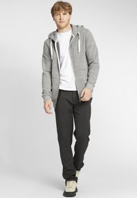 Sivi zip-up dukserica s kapuljačom, u paru s tamnim hlačama i svijetlim obućom. Glatka tekstura, ležeran dizajn i bočni džepovi.