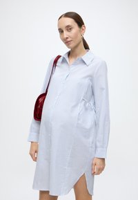 Lichtblauwe gestreepte zwangerschapsblousejurk met knopen aan de voorkant, zijstrikjes en lange mouwen. Gecombineerd met een dieprode handtas.